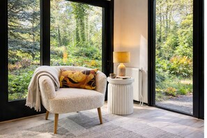 Une pièce confortable avec un canapé, une lampe et une vue sur une forêt, visible à travers de grandes fenêtres.