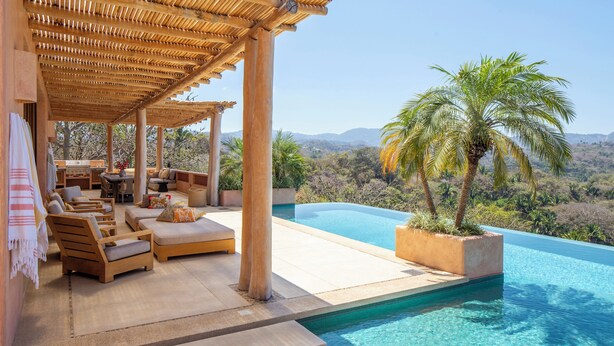 Zona junto a la piscina con tumbonas de madera, un pérgola y vistas a una cordillera.