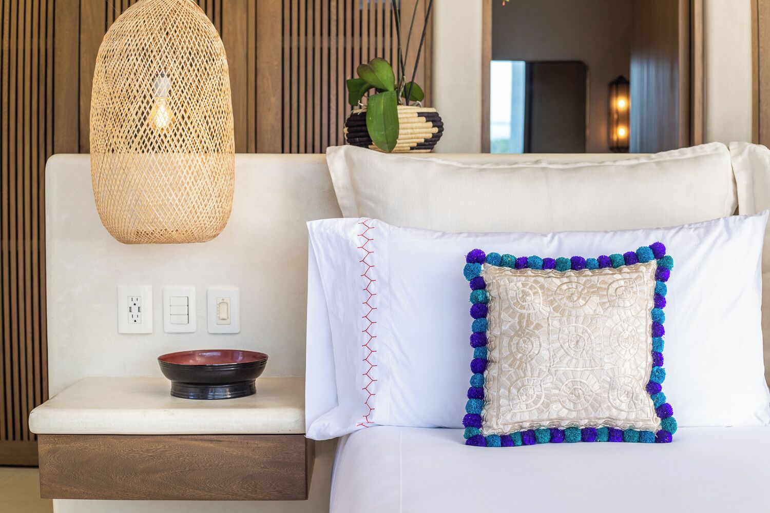Een moderne hotelkamer met een witte bank, een decoratieve geweven lamp, een nachtkastje met een rode kom en een decoratief kussen met blauwe en paarse pompons.
