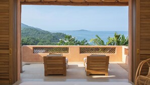 Un balcón con dos sillas de madera y una de mimbre, con vistas a un paisaje costero con vegetación y el mar.