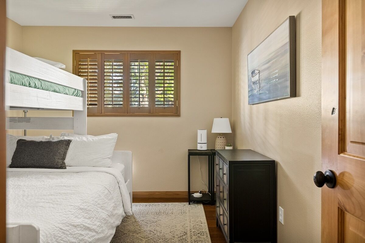 Un dormitorio con una cama litera, una mesita de noche con lámpara, un aparador y una ventana con persianas.