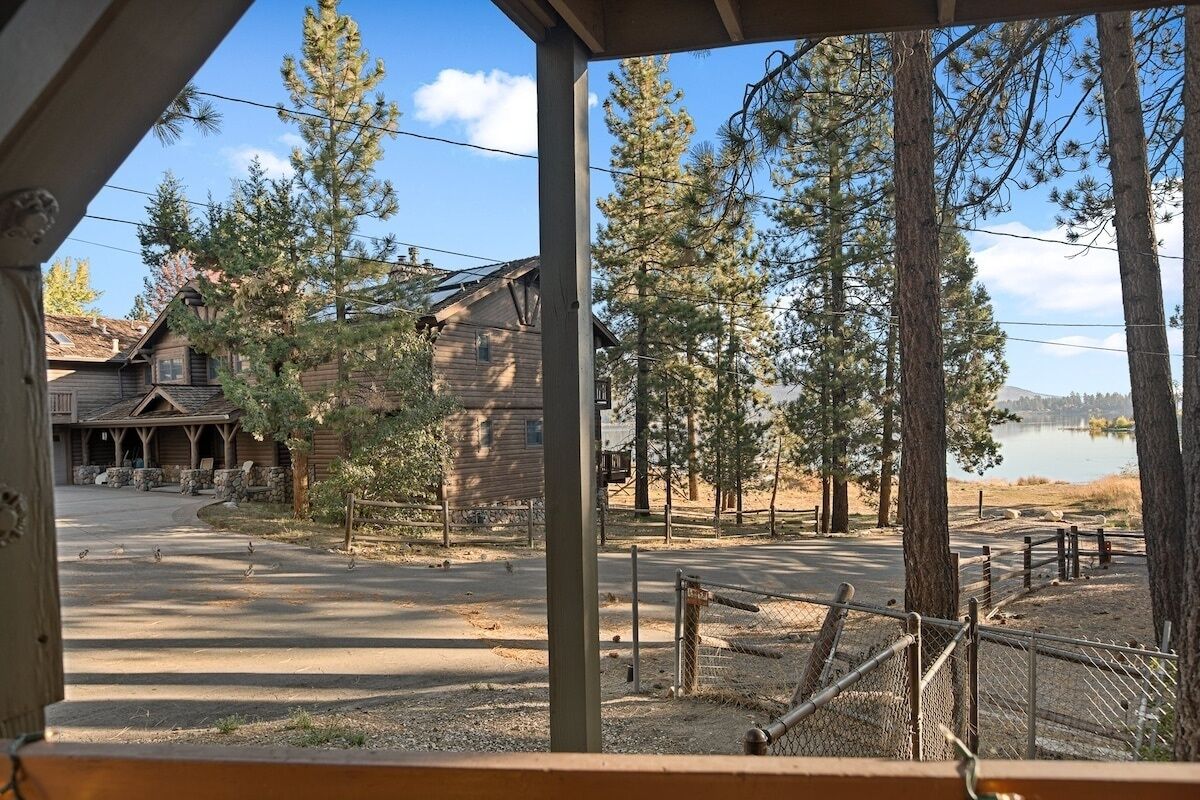 A rustic lodge with a wooden exterior, a paved driveway, and a view of a lake surrounded by tall pine trees.