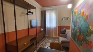 A compact room with a bed, a desk, a chair, a TV, and a painting of cacti. - Capital Central Suites No3 - Cikka (Valletta, Malta)