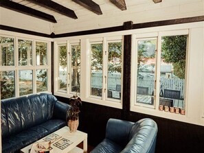 A room with a black leather sofa, a small table with a plant, and large windows with a view of a fenced yard. - Splendid Cabin in Camping Resort (Graested)