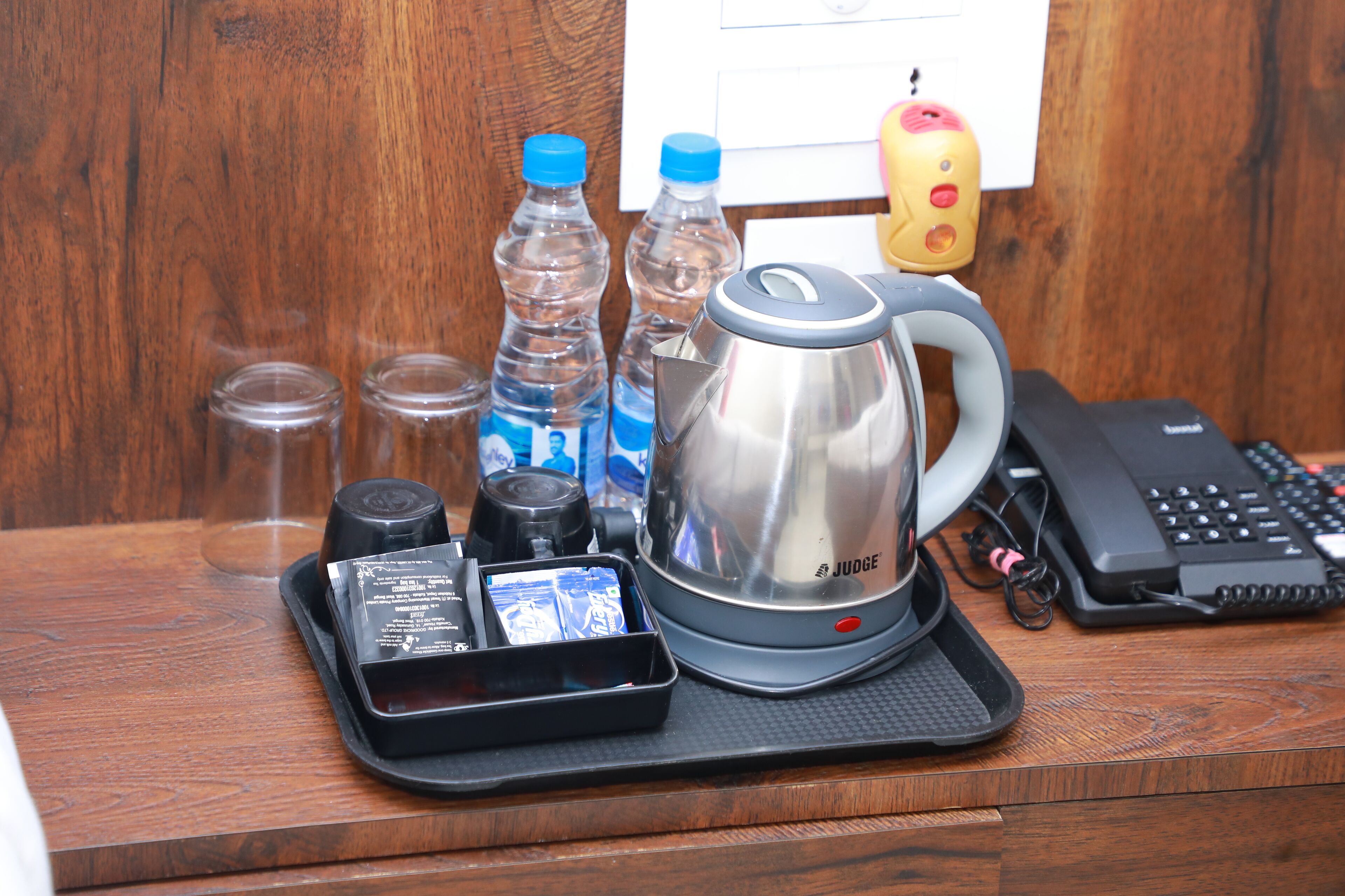Een houten bureau met een koffiezetapparaat, twee waterflessen, een telefoon en een dienblad met een klein doosje en een kop.