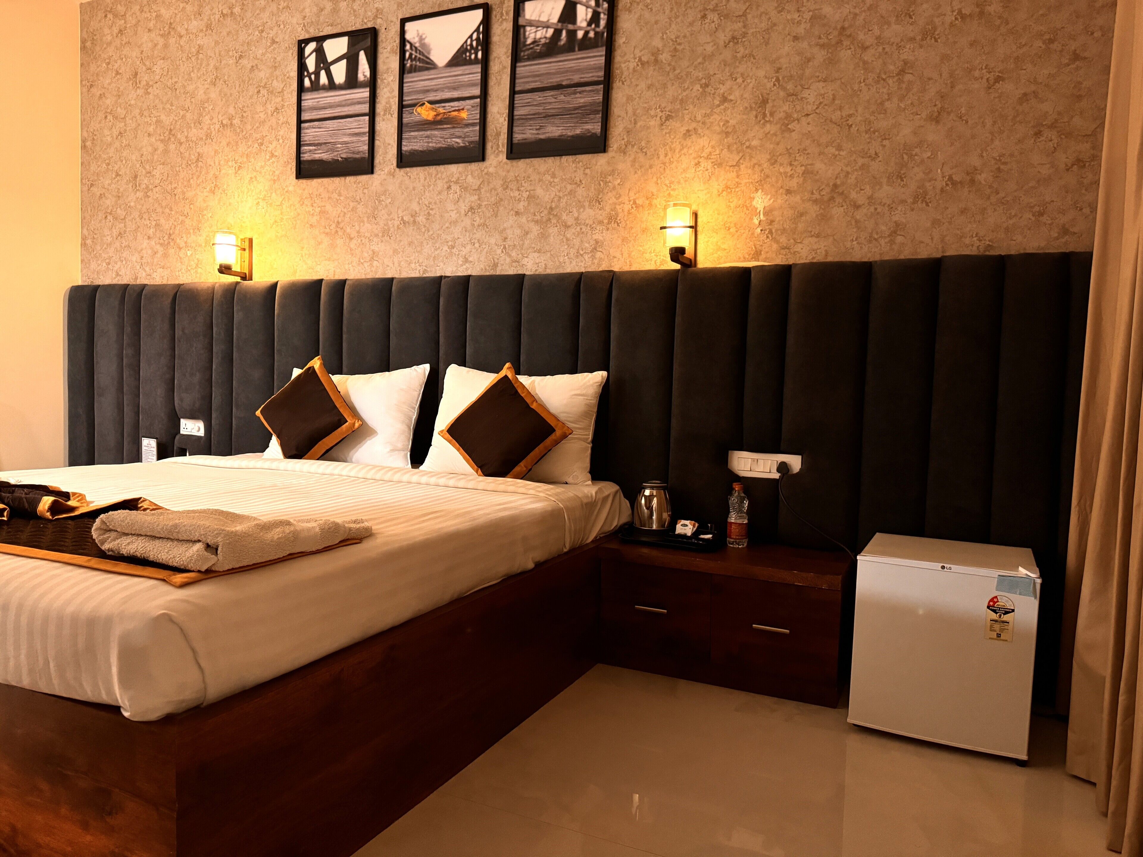 A hotel room with a large bed, a nightstand, a small refrigerator, and wall-mounted artwork.