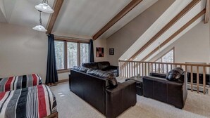 Un salon de style industriel, avec un plafond haut, des poutres apparentes en bois, un ensemble de canapés en cuir noir, un canapé à rayures et un escalier menant à un niveau supérieur.