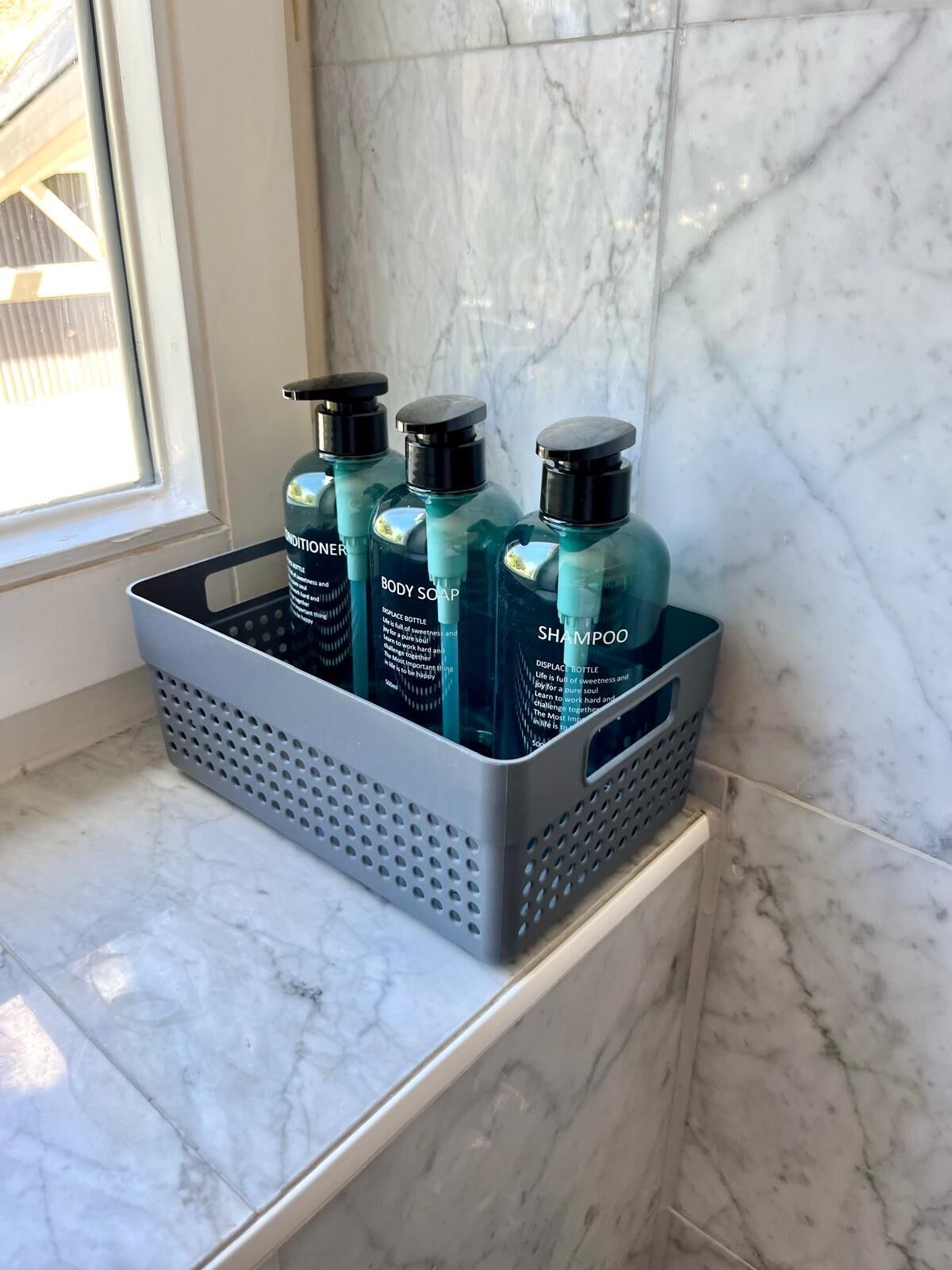 Une salle de bain avec un sol en marbre et une fenêtre. On y trouve un panier gris contenant trois bouteilles de savon pour le corps et de shampoing.