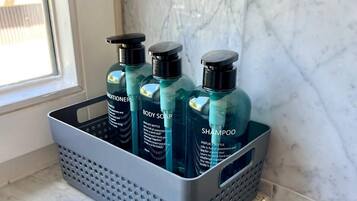 Une salle de bain avec un sol en marbre et une fenêtre. On y trouve un panier gris contenant trois bouteilles de savon pour le corps et de shampoing.
