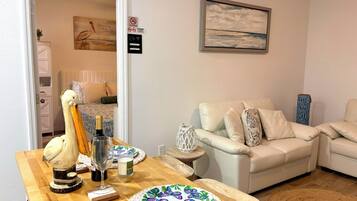 A room with a wooden table set with a bottle, glass, candle, and a decorative bird. There is a white sofa with cushions and a blue patterned pillow. A framed painting hangs on the wall, and a 'No Smoking' sign is visible.