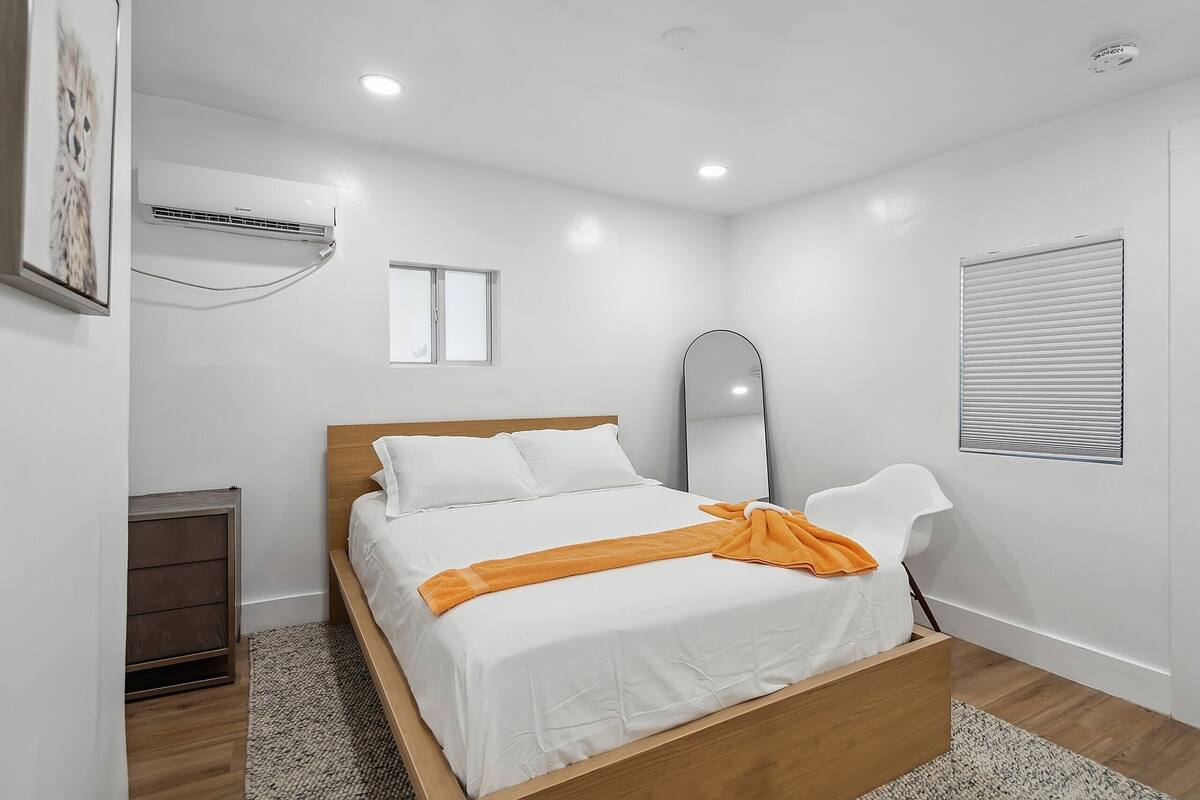 A modern bedroom with a wooden bed, white bedding, a large mirror, and a window with blinds.