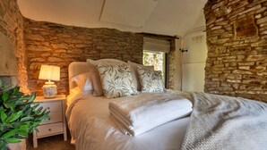 Un dormitorio con pared de piedra, una cama con edredón blanco y almohadas estampadas, una mesita de noche con lámpara y una planta en maceta.
