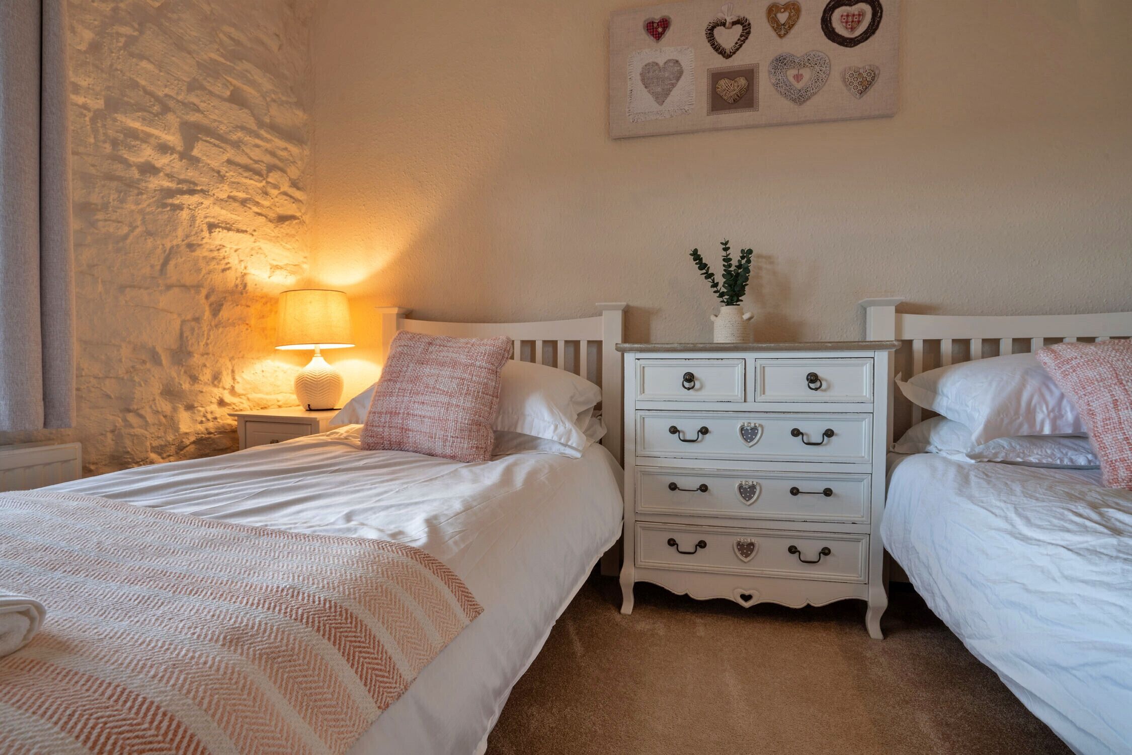 Une chambre avec deux lits, un mur en pierre, une table de chevet avec une lampe et une commode avec des poignées en forme de cœur.