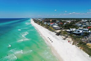 Luftaufnahme einer Küstenstadt mit einem weißen Sandstrand, klarem türkisblauem Wasser und einer Mischung aus Wohn- und Geschäftsgebäuden.
