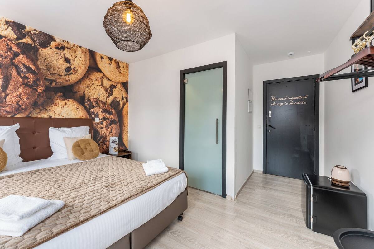 A modern hotel room with a large bed, a wall mural of cookies, a dark door with a chocolate-themed sign, and a small kitchenette.