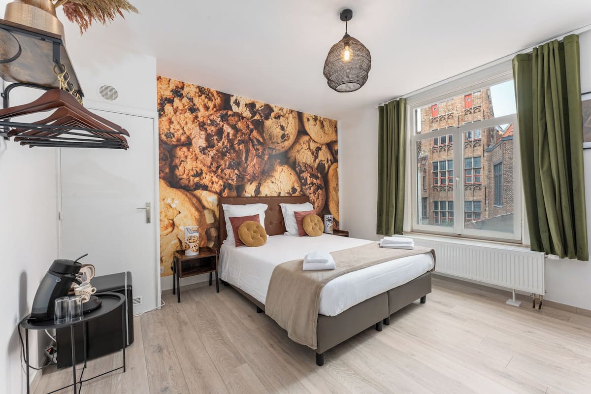 A modern bedroom with a large bed, a coffee machine, a nightstand, a wall-mounted shelf, and a window with curtains.