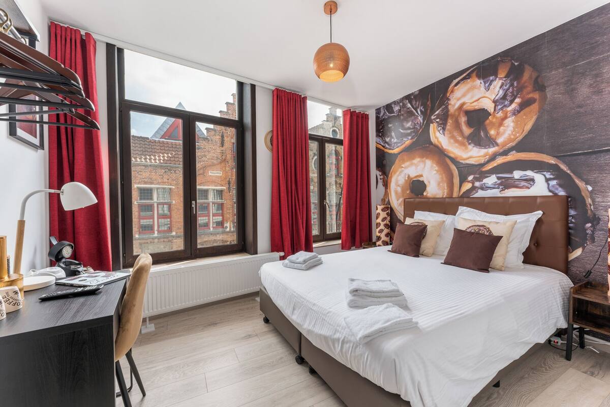 A modern hotel room with a large bed, red curtains, a city view, and a coffee-themed wall mural.