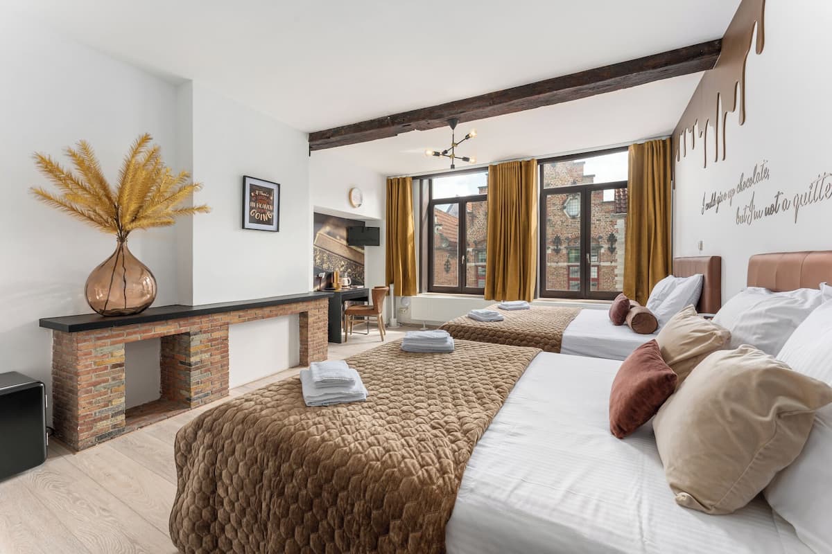 A hotel room with a brick fireplace, two beds with brown and white bedding, a large window with curtains, and a view of a brick building outside.