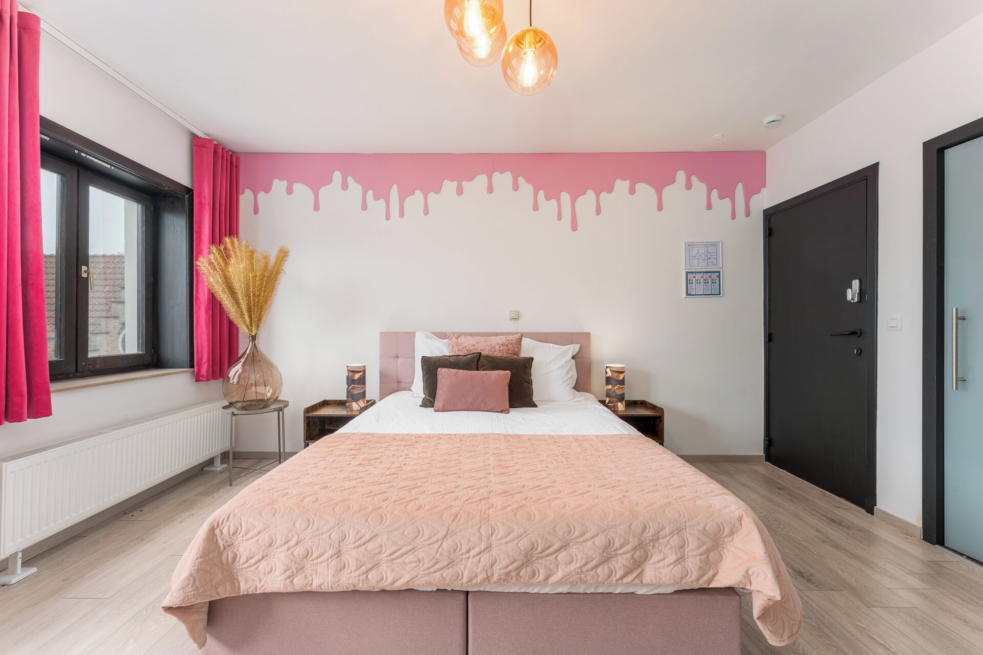 Un dormitorio con una cama grande, una ventana con cortinas rojas, una pared con diseño de pintura goteando y una puerta.