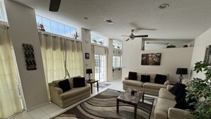 A spacious living room with two sofas, a coffee table, and a large rug. There is a ceiling fan, a painting on the wall, and a plant in the corner. - Villa Crayson (Kissimmee)