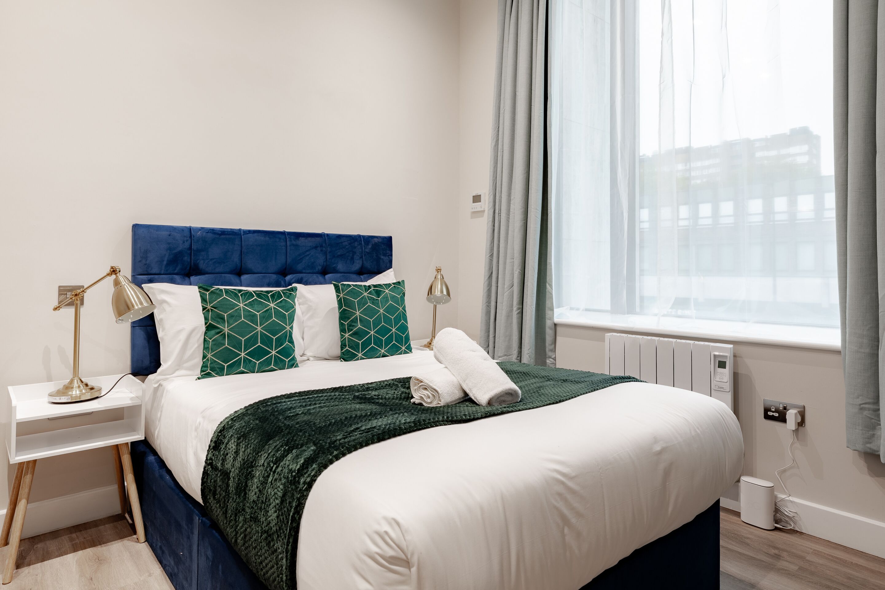 A neatly made bed with a blue headboard, white bedding, and green patterned pillows. There is a bedside table with a lamp, a window with curtains, and a view of buildings outside.
