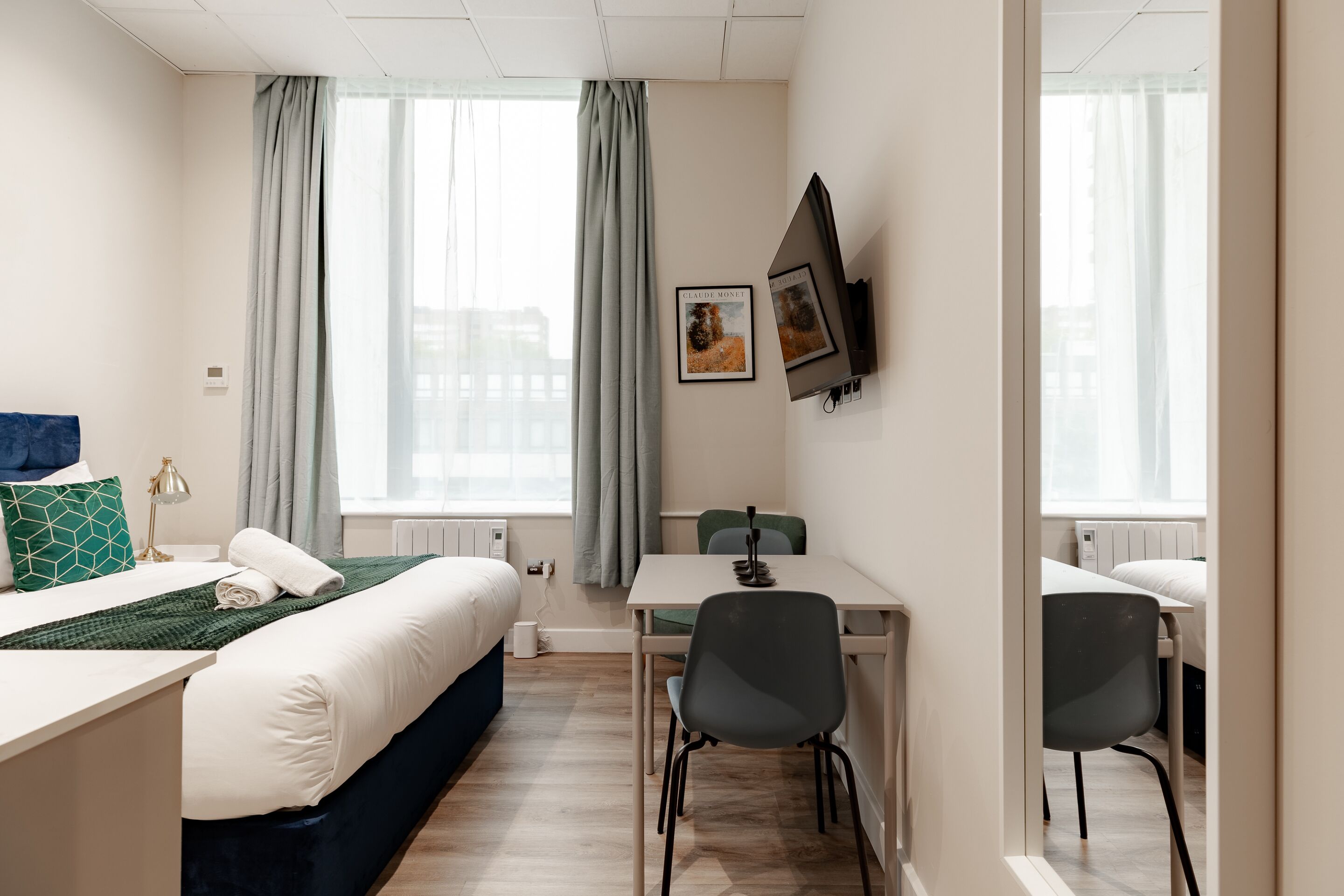 Une chambre d’hôtel avec un lit, un bureau, deux chaises, une télévision et une fenêtre avec des rideaux.