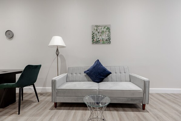 Una sala de estar moderna con un sofá gris, un sillón verde, una mesa de centro de vidrio, una lámpara de pie y un reloj de pared.