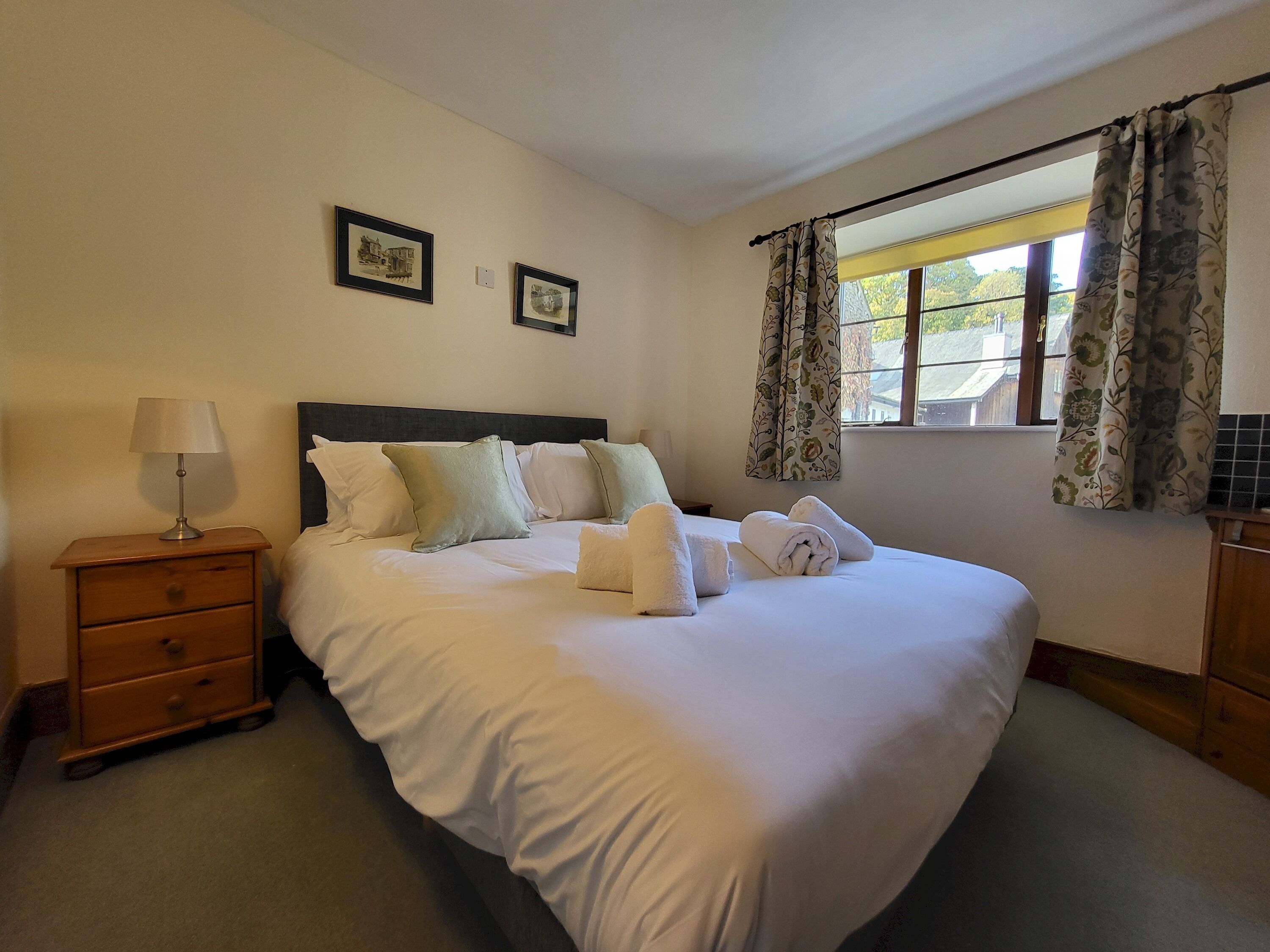 A neatly made bed with white linens and pillows, a bedside table with a lamp, and two framed pictures on the wall.