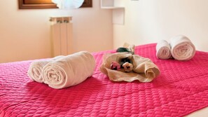 A bed with a pink quilted bedspread, two rolled towels, and a small basket with a towel and a pair of slippers.