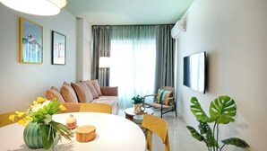 A modern living room with a sofa, a dining table with chairs, a potted plant, and a flat-screen TV mounted on the wall.