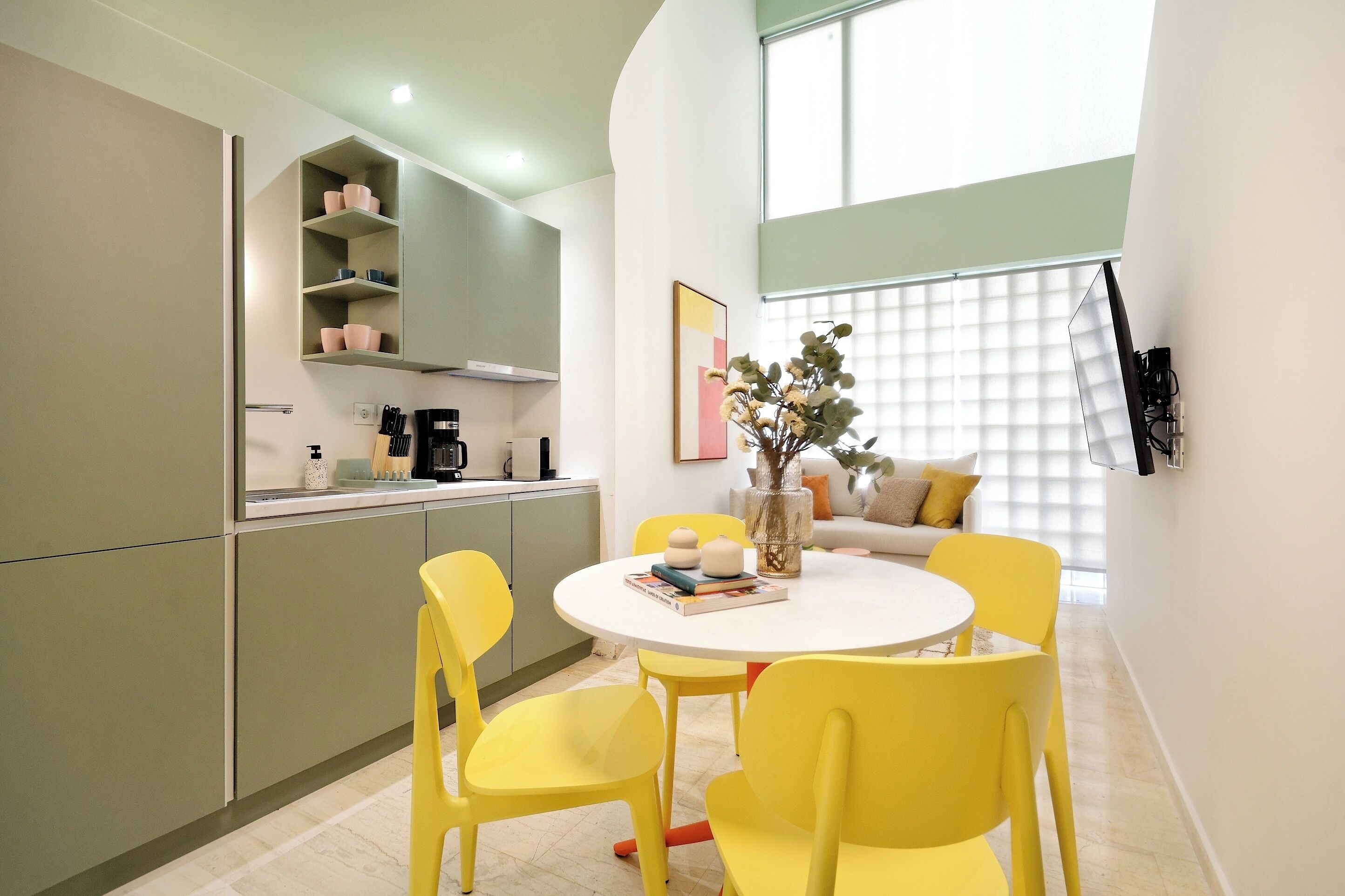 A modern kitchen with green cabinets, a white countertop, and a dining area with yellow chairs and a white table.