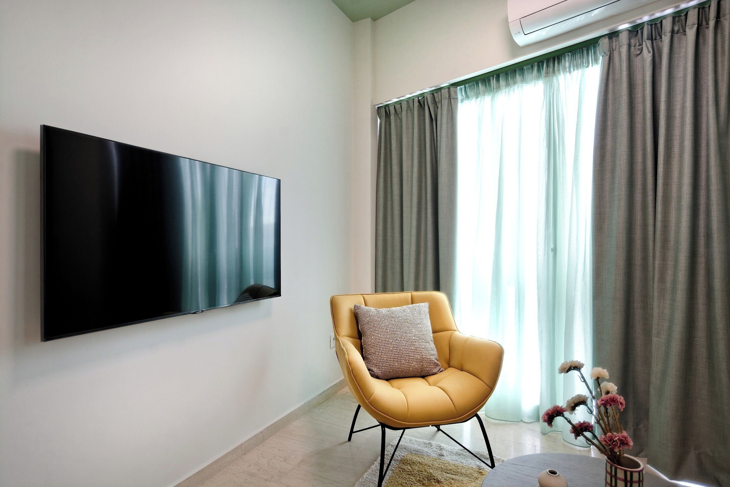 Un salon moderne avec un téléviseur à écran plat fixé au mur, un fauteuil jaune agrémenté d’un coussin et une grande fenêtre ornée de rideaux gris.
