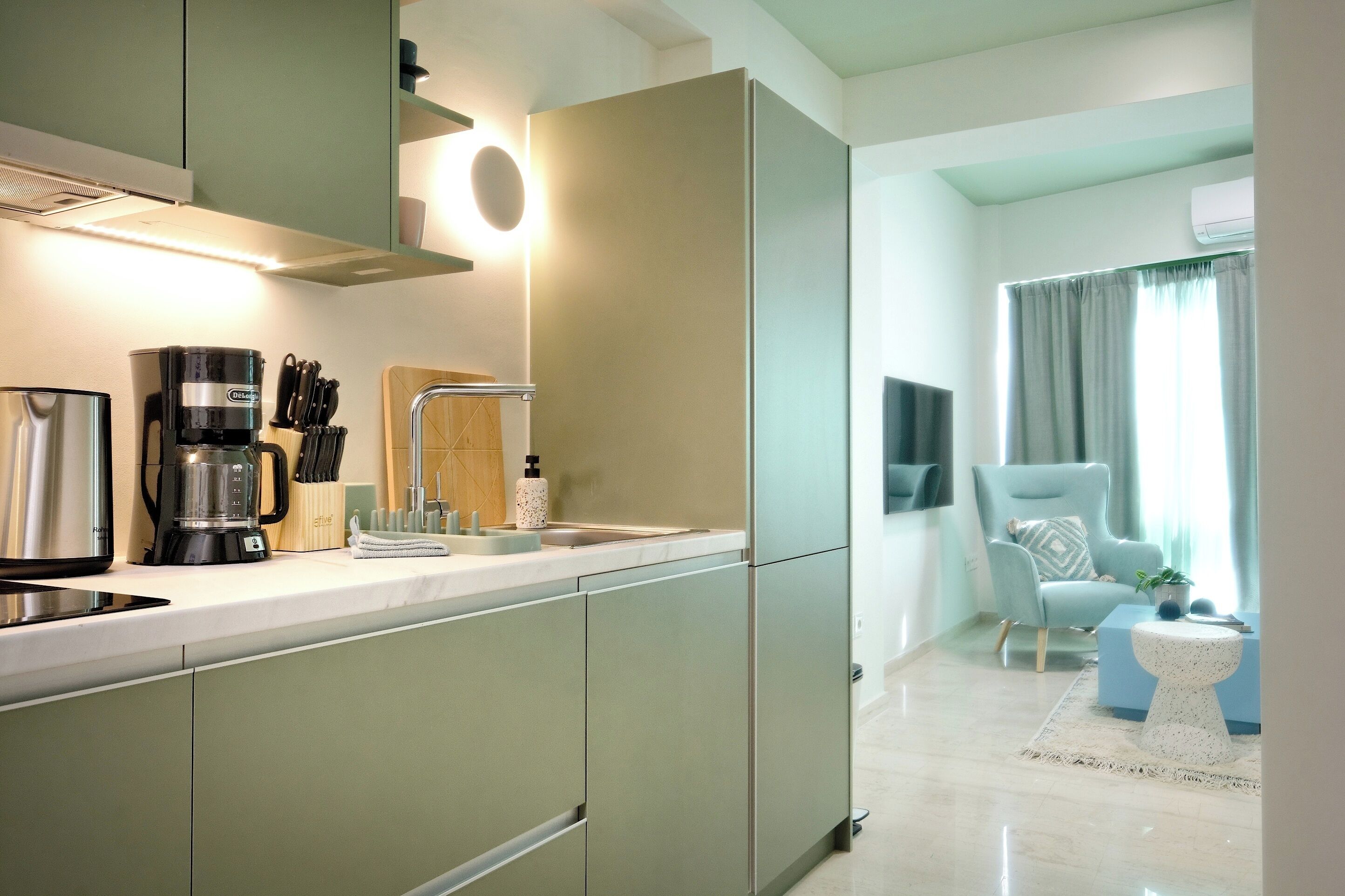 A modern kitchen with a coffee maker, knives, and a sink. There is a chair and a small table in the adjacent living area.