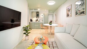 A modern living room with a white sofa, a round orange coffee table, and a dining area with a white table and pink chairs. There is a kitchen in the background with a white countertop and a refrigerator.