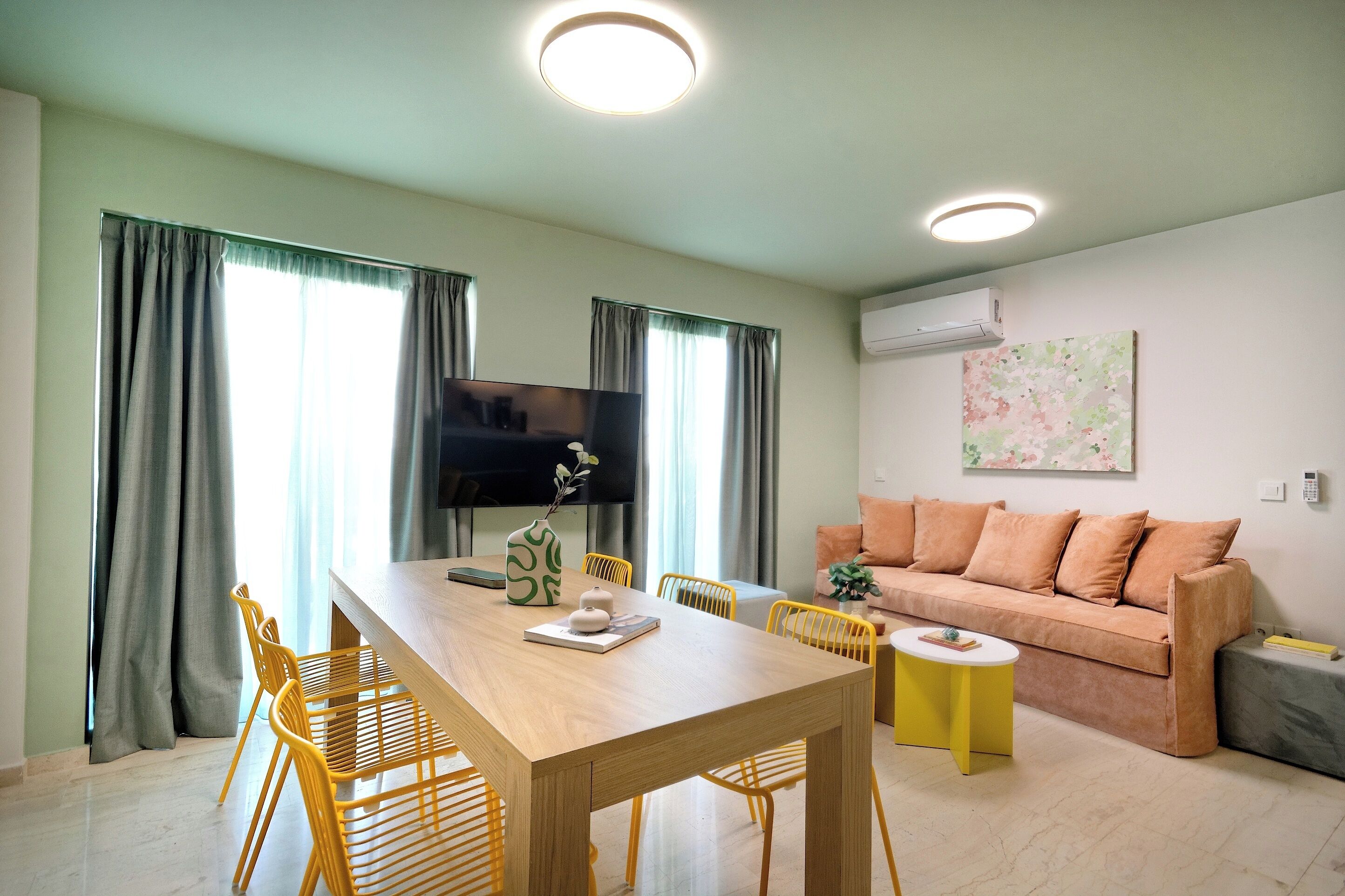 A modern living room with a wooden dining table, yellow chairs, a sofa, and a flat-screen TV.