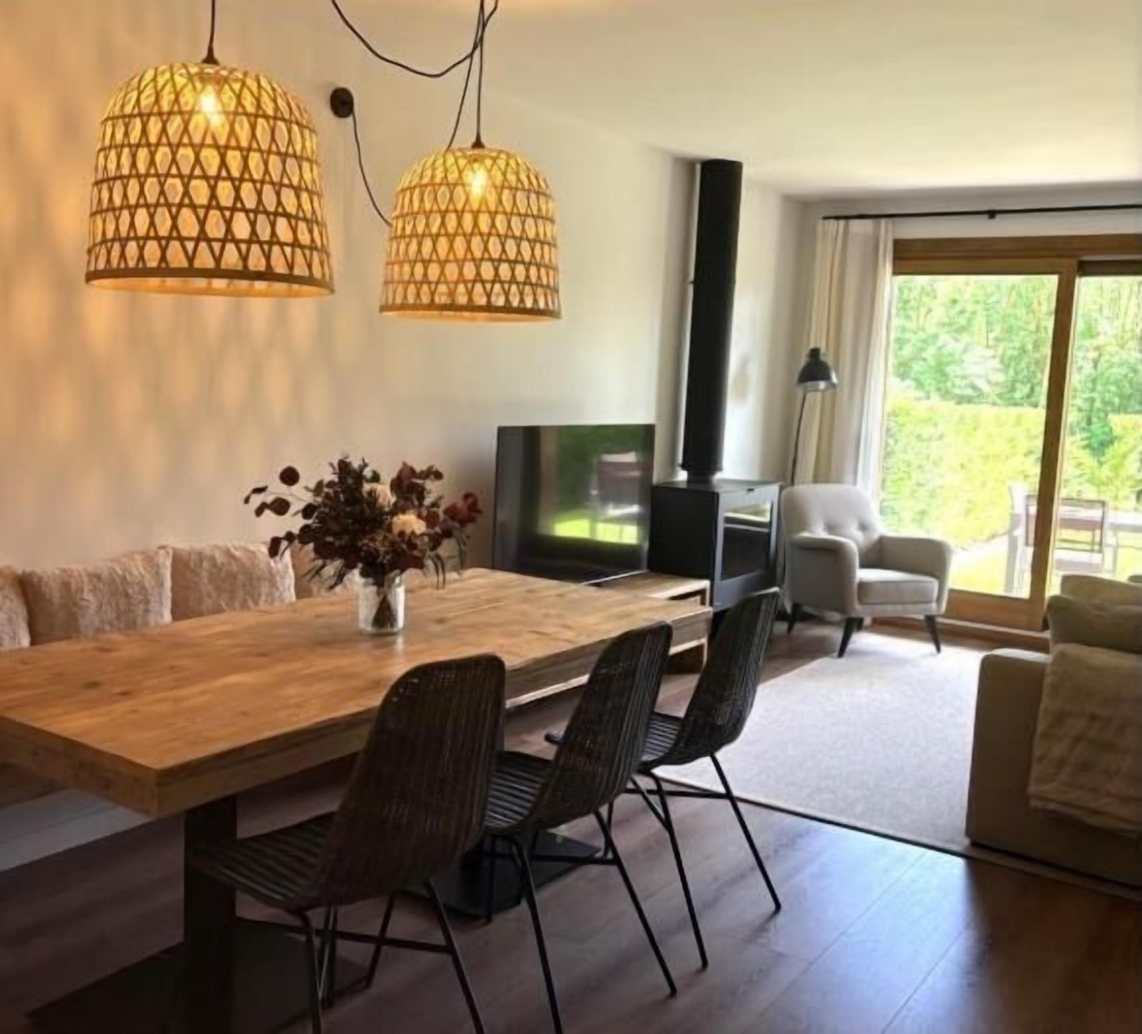 Een moderne woonkamer met een houten eettafel, zwarte stoelen, een televisie en een bank. Er hangen twee lampen met een geometrisch patroon.