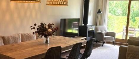 Een moderne woonkamer met een houten eettafel, zwarte stoelen, een televisie en een bank. Er hangen twee lampen met een geometrisch patroon.