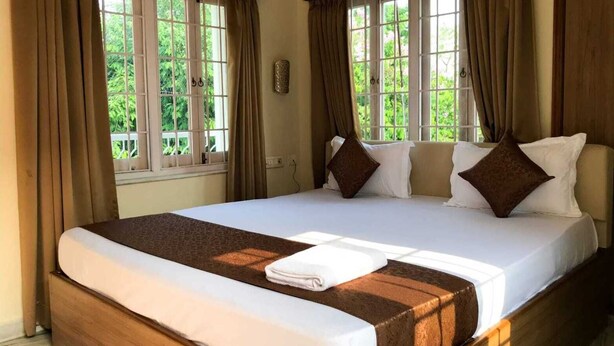 A bedroom with a large bed, two windows with curtains, and a view of greenery outside. - Ashray The Living Entity  (Bidhannagar)