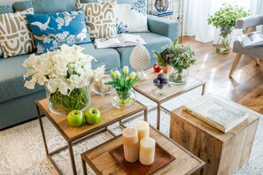 Un salon avec un canapé bleu, des coussins décoratifs, une table basse avec des pommes et des bougies, et un vase rempli de fleurs blanches.