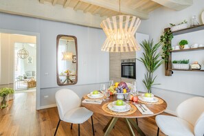 Un coin repas avec une table ronde dressée pour quatre personnes, entourée de chaises blanches. Un grand miroir est accroché au mur, et un plafond en bois avec des poutres apparentes est visible.