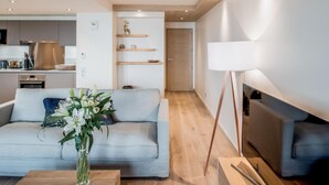 Una sala moderna con un sofá gris, una mesa de centro y un jarrón de flores. Se ve una cocina al fondo, con estufa y armarios.