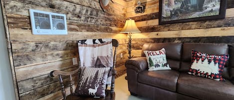 A cabin interior with a wooden wall, a leather couch, a wooden chair, and a lamp.