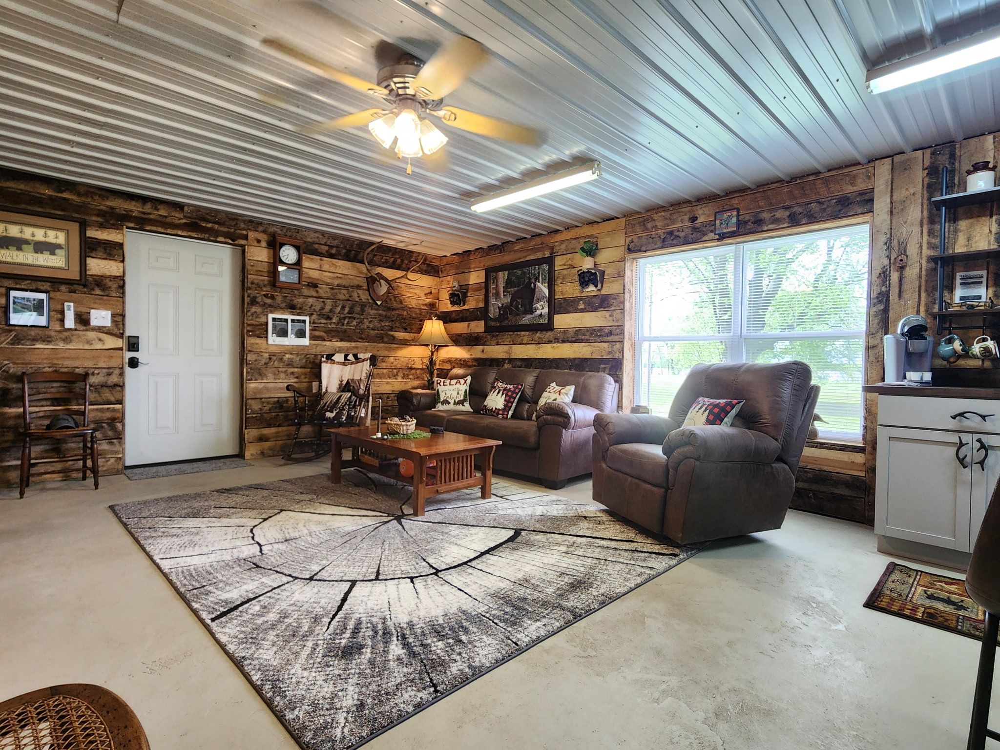 A rustic interior with a wooden wall, a ceiling fan, a fireplace, and a large window.