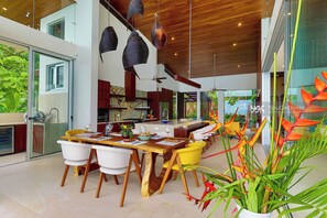 Un comedor moderno con una mesa y sillas de madera, una cocina al fondo y un amplio ventanal con vistas a la vegetación.