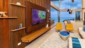Een moderne woonkamer met een grote televisie, houten lambrisering, gele stoelen en uitzicht op zee.