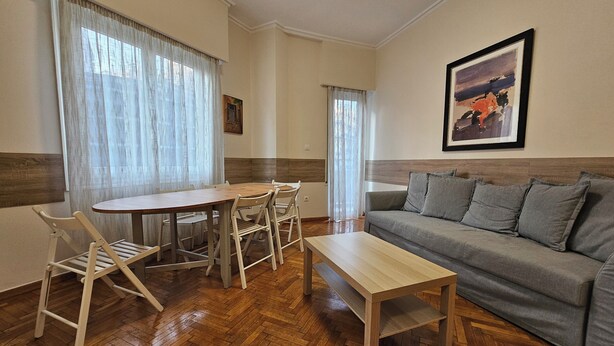 Een woonkamer met een houten visgraatvloer, een grijze bank, een houten salontafel en een eettafel met vier stoelen.