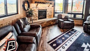 A rustic interior with a stone fireplace, leather sofas, and large windows offering a view of the outdoors.