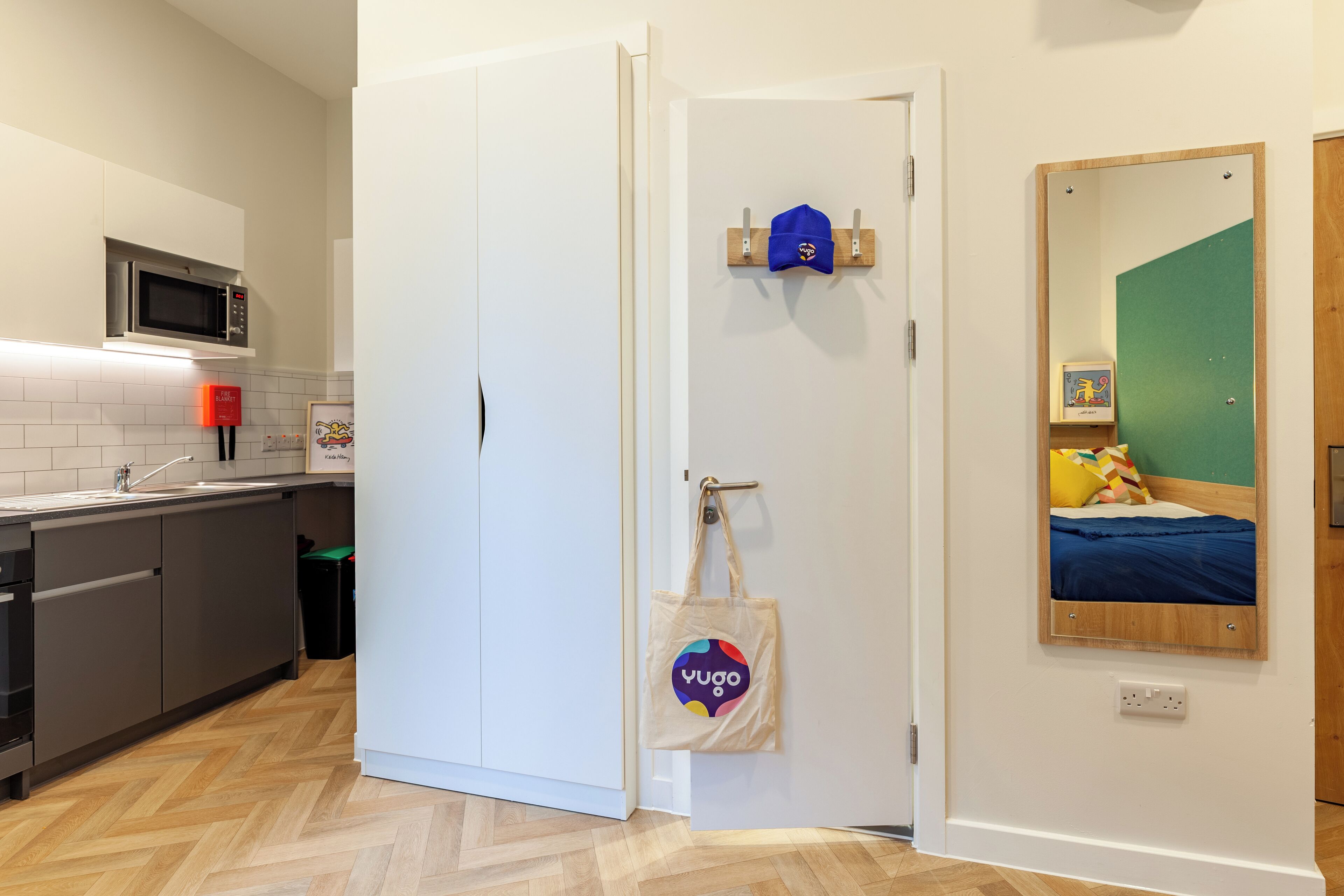 A modern kitchen with a microwave, a white door, and a mirror reflecting a bed with colorful pillows.