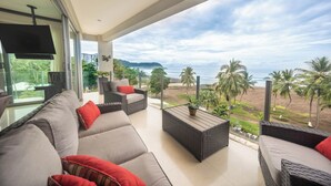 En moderne stue med en stor sofa, et sofabord og et fjernsyn. Stuen har et stort vindue med udsigt over en strand og palmer.