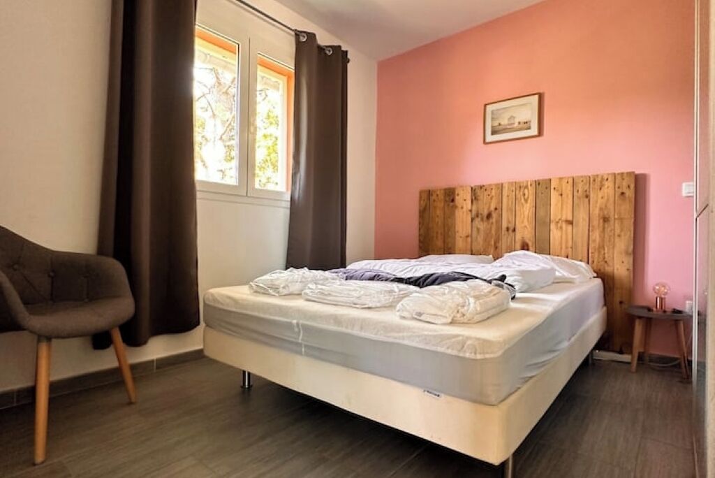 Une chambre à coucher avec une tête de lit en bois, un lit recouvert de draps blancs, un fauteuil marron et une fenêtre avec des rideaux.